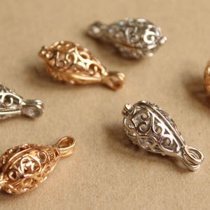May include: Close-up of several filigree teardrop-shaped jewelry components in gold and silver tones. Each component has an openwork design and a small loop at the base. The components are scattered on a neutral background.