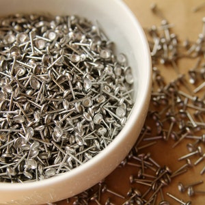 May include: A close-up shot of a white bowl filled with small, silver upholstery tacks. The tacks have a round, flat head and a slender, pointed shank. Some tacks are scattered outside the bowl on a light brown surface.