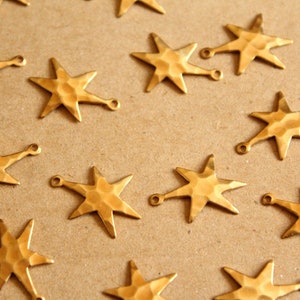 May include: A collection of 14 gold-toned metal star charms with a hammered texture. Each star has a small loop at the top for attaching to jewelry or other crafts.