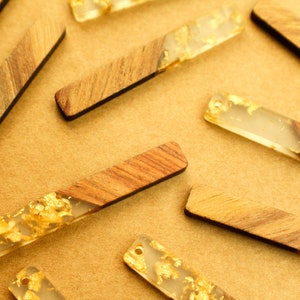 May include: Close-up of several rectangular wooden pendants with a section of clear resin embedded with gold leaf. The wood has a natural grain, and the resin section is near the end of each pendant. The pendants are scattered on a tan surface.