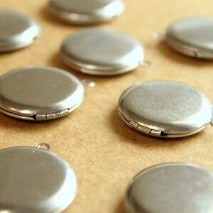 May include: A collection of ten silver-toned locket pendants with a hinged closure. The lockets are arranged on a brown surface.