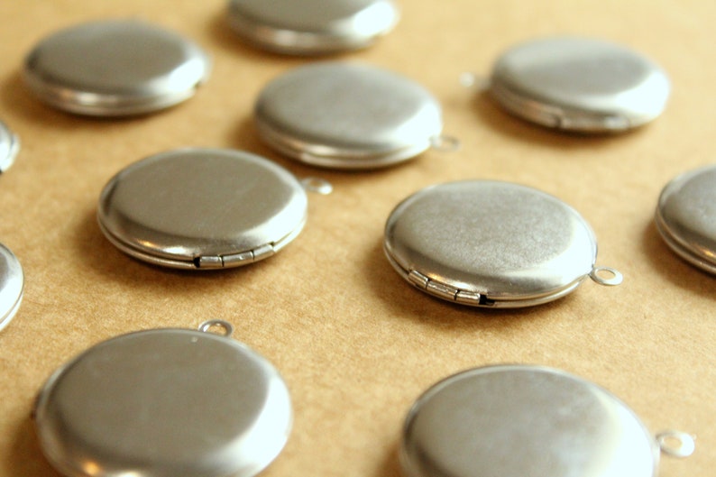 May include: A collection of ten silver-toned locket pendants with a hinged closure. The lockets are arranged on a brown surface.