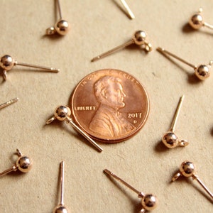 May include: A close-up of a group of 14 gold-toned earring findings with a small ball at the end of each post. The findings are arranged around a US penny.