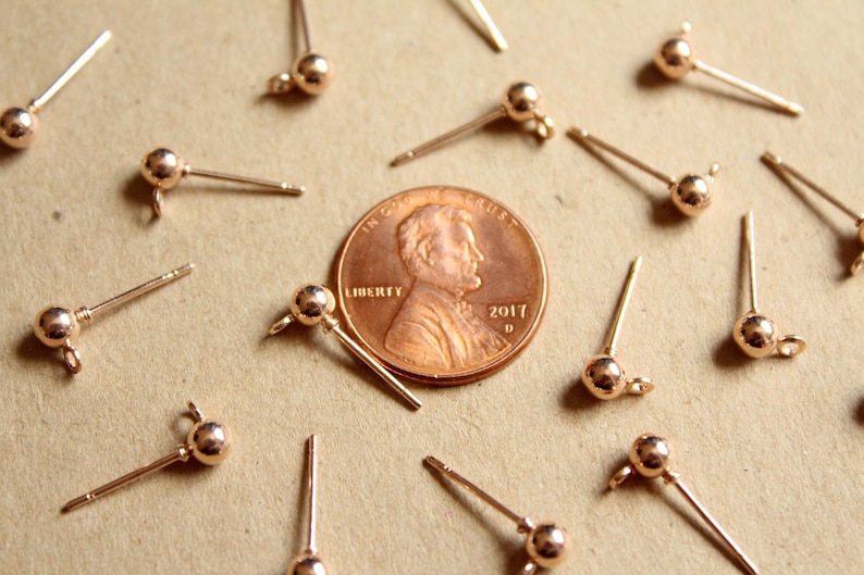 May include: A close-up of a group of 14 gold-toned earring findings with a small ball at the end of each post. The findings are arranged around a US penny.