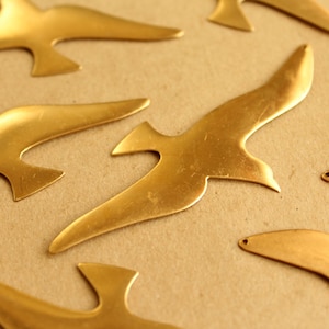 May include: A collection of seven gold-colored metal bird cutouts. The birds are in various sizes and shapes, with some having a more realistic bird shape and others being more abstract.