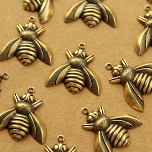 Könnte beinhalten: Eine Sammlung von 13 goldfarbenen Metall-Bienenanhängern mit detaillierten Flügeln und segmentierten Körpern. Die Anhänger sind auf einer braunen Oberfläche verstreut angeordnet.