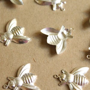 Puede incluir: Una colecci&oacute;n de abalorios de abeja plateados con alas detalladas y cuerpos texturizados. Los abalorios est&aacute;n esparcidos sobre una superficie marr&oacute;n.