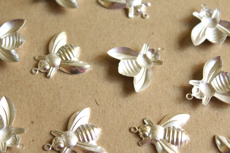 Puede incluir: Una colecci&oacute;n de abalorios de abeja plateados con alas detalladas y cuerpos texturizados. Los abalorios est&aacute;n esparcidos sobre una superficie marr&oacute;n.