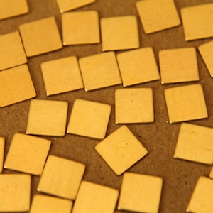 Op de afbeelding: Een close-up van een verzameling kleine, vierkante, goudkleurige metalen tegels die verspreid liggen op een bruin oppervlak.