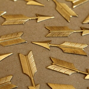 May include: A collection of gold-coloured arrow-shaped metal embellishments. The arrows are arranged in a random pattern on a brown surface.