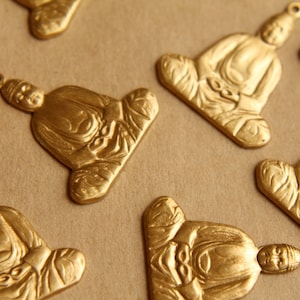 May include: A collection of ten gold-colored metal charms featuring a seated Buddha figure. Each charm is intricately detailed and has a slightly different design.