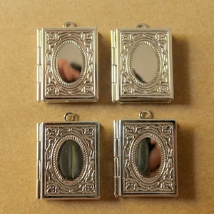 May include: Four small, silver-colored book-shaped pendants. Each pendant has an oval frame with a decorative border. The pendants are hinged and have a small loop at the top for attaching to a chain or other jewelry.