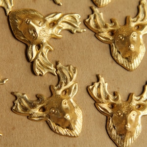 May include: Close-up of several gold-colored deer head embellishments. Each deer head has detailed antlers and facial features. The embellishments are arranged on a tan surface, creating a repeating pattern. These are likely for crafting or decorative purposes.