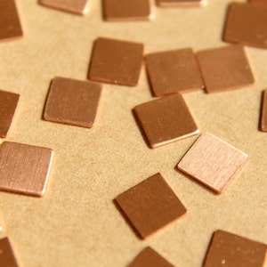 May include: Close-up shot of numerous small, square copper metal pieces scattered on a light brown surface. The squares have a slightly reflective surface and vary in shade from light to dark copper.