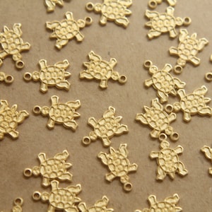 May include: A collection of gold-toned turtle charms, each with a detailed textured shell design. The charms are scattered on a light brown surface.