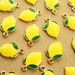 May include: Close-up of multiple yellow lemon charms with green leaves and gold-colored metal accents. Each charm has a small loop for attaching to jewelry or other crafts. The charms are scattered on a light brown surface.