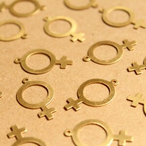 May include: A close-up of a group of gold-colored metal charms shaped like the female gender symbol. The charms are scattered on a brown surface.