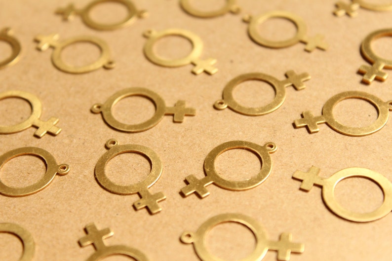 May include: A close-up of a group of gold-colored metal charms shaped like the female gender symbol. The charms are scattered on a brown surface.