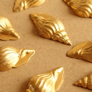 May include: A collection of seven gold-colored seashell-shaped decorative ornaments. The shells are arranged on a brown surface.