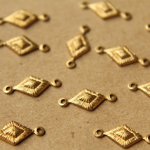 Puede incluir: Una colección de conectores de metal de forma de diamante de color dorado con dos bucles a cada lado. Los conectores están dispersos sobre una superficie marrón.
