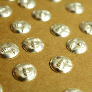 May include: A close-up of a collection of silver-toned metal buttons with a moon face design. The buttons are arranged in rows on a brown surface.
