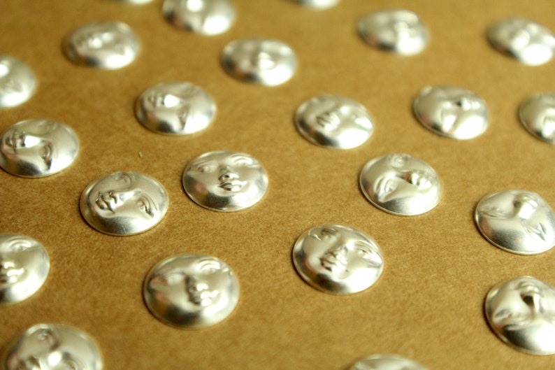 May include: A close-up of a collection of silver-toned metal buttons with a moon face design. The buttons are arranged in rows on a brown surface.