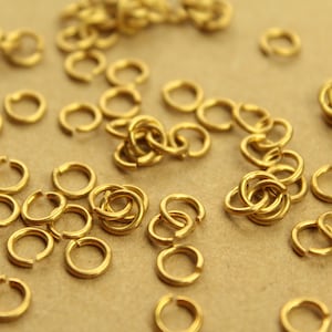 Könnte beinhalten: Nahaufnahme einer Sammlung kleiner, offener Sprungringe in warmem Goldton. Diese Metallringe sind auf einer neutralen Oberfläche verstreut, wobei einige Ringe zusammengefasst und andere verteilt sind. Ideal für Schmuckherstellung und Bastelarbeiten.