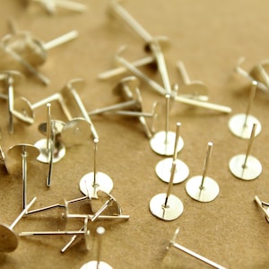 May include: A close-up of a pile of silver earring posts with flat backs. The posts are scattered on a brown surface.