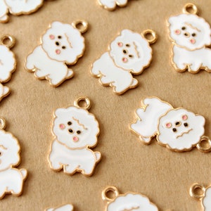 May include: White and gold-colored Bichon Frise dog charms. Each charm features a white dog with a gold outline and a small loop for attaching to jewelry or other crafts. The dogs have black eyes and pink cheeks.