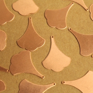 May include: Close-up of multiple copper-colored metal charms. Each charm has a unique ginkgo leaf shape with a small hole at the top for stringing. The charms are scattered on a textured, olive-green surface.
