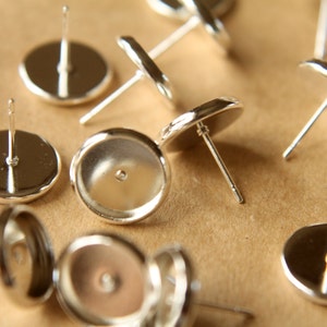 May include: Silver earring settings with a round, flat base and a post for attaching earrings. The settings are designed to hold a variety of cabochons or other decorative elements.