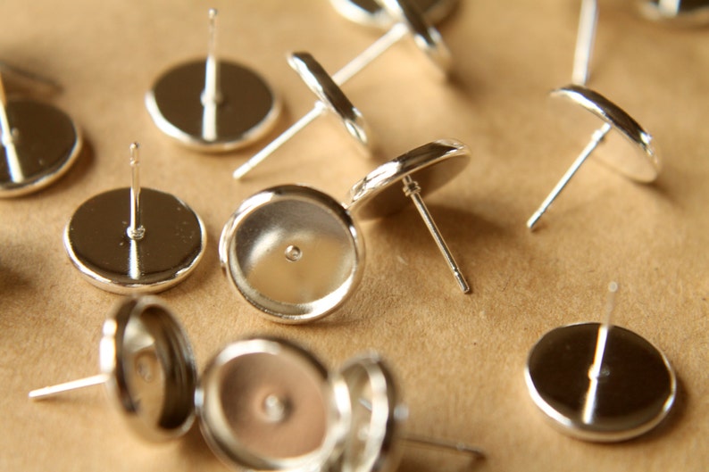 May include: Silver earring settings with a round, flat base and a post for attaching earrings. The settings are designed to hold a variety of cabochons or other decorative elements.