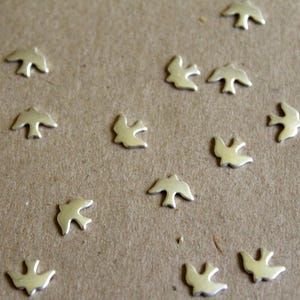 Peut inclure: Petits ornements dorés en forme d'oiseaux dispersés sur une surface marron. Les oiseaux sont orientés dans différentes directions, les ailes déployées. Ces éléments décoratifs pourraient être utilisés pour l'artisanat ou la bijouterie.