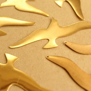 May include: A collection of gold-colored metal bird cutouts on a brown background. The birds are all different sizes and shapes, and some have a slightly textured surface.