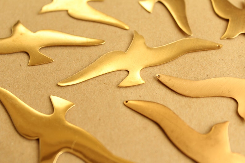 May include: A collection of gold-colored metal bird cutouts on a brown background. The birds are all different sizes and shapes, and some have a slightly textured surface.