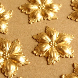 May include: A collection of six gold-toned metal flower-shaped embellishments with intricate details. The embellishments are arranged on a brown surface.