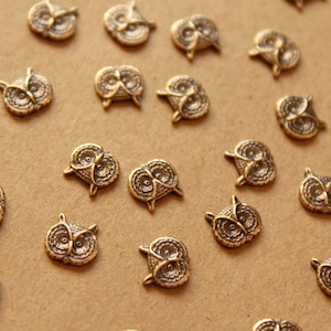 May include: A collection of gold-toned metal owl charms with detailed features. The charms are scattered on a brown surface.