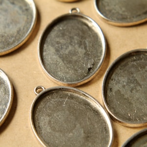 May include: Close-up of several silver-toned oval pendant trays with small loops for attaching chains. The trays have a textured, gray interior and a shiny silver border. The pendants are arranged on a light brown surface.