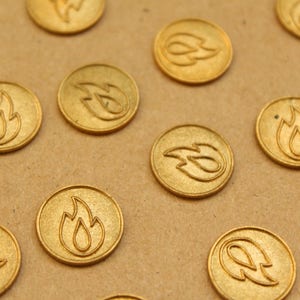 Puede incluir: Una colección de sellos de cera dorados con un diseño de llama. Los sellos están dispuestos en un patrón sobre una superficie marrón.