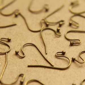 May include: A close-up of a group of gold-toned earring hooks. The hooks are made of metal and have a small loop at the top. The hooks are scattered on a brown surface.