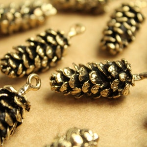 Może przedstawiać: Zbliżenie kilku małych, brązowych zawieszek w kształcie szyszki. Każda zawieszka ma szczegółową, teksturowaną powierzchnię i pętlę na górze do mocowania do biżuterii lub innych rękodzieł. Zawieszki są rozrzucone na jasnobrązowej powierzchni.