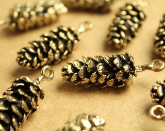 May include: A collection of small, bronze-coloured pinecone charms. Each charm has a detailed, textured surface and a loop at the top for attaching to jewellery or other crafts. The charms are scattered on a light brown surface.