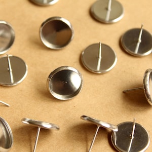 May include: A close-up of a group of silver earring settings. The settings are round and have a small post on the back. They are arranged on a brown surface.