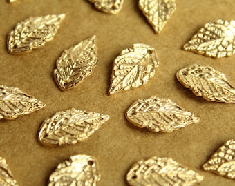 Puede incluir: Una colección de abalorios de hojas de metal de color dorado. Las hojas tienen un diseño detallado en relieve y están esparcidas sobre una superficie marrón.