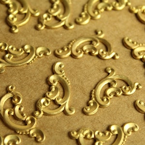 May include: A collection of gold-coloured metal decorative embellishments with a scroll design. The embellishments are arranged on a brown surface.