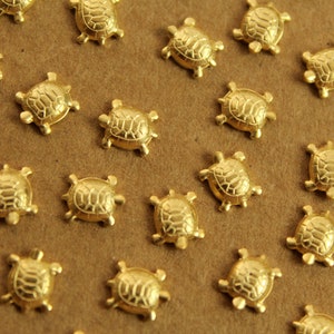 May include: A collection of small, gold-colored turtle charms scattered on a brown surface. Each charm has a detailed shell design and four small legs, creating a whimsical and nature-inspired aesthetic. These charms are ideal for jewelry making or craft projects.