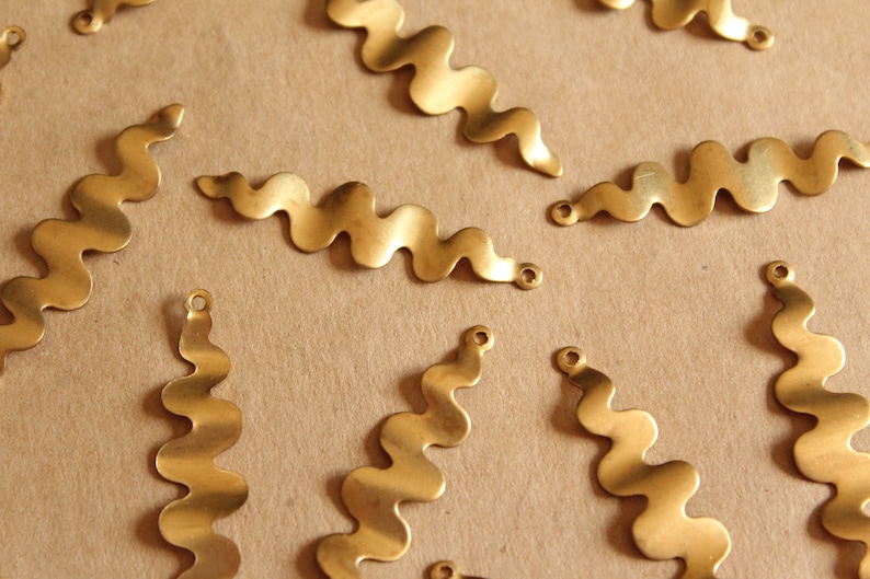 K&ouml;nnte beinhalten: Eine Sammlung von wellenf&ouml;rmigen, goldfarbenen Metallformen mit kleinen L&ouml;chern an den Enden. Die Formen sind auf einer braunen Oberfl&auml;che verstreut.