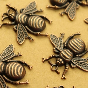 May include: Close-up of several bronze-colored bee charms. Each charm features detailed wings, a striped abdomen, and delicate legs and antennae. The charms are scattered on a light brown surface, showcasing their intricate design.
