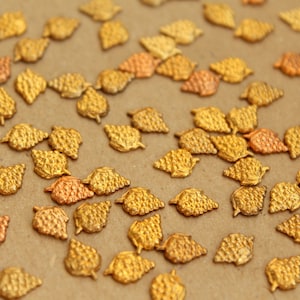 May include: A large quantity of small, gold-coloured metal leaves scattered on a brown surface. The leaves have a textured, raised design.