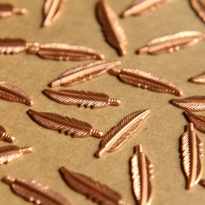 May include: A collection of copper-colored feather charms scattered on a brown surface. The feathers are all the same size and shape, and they have a delicate, intricate design.